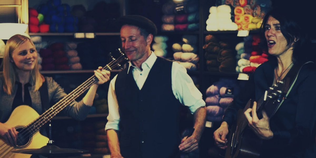 A man and two women with guitarrs in front of a shelf full of yarn. 