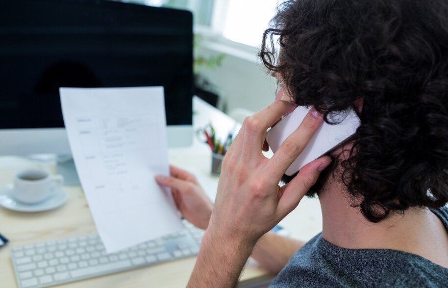 Mann am Schreibtisch telefoniert und hält ein Call Sheet vor einem Computerbildschirm
