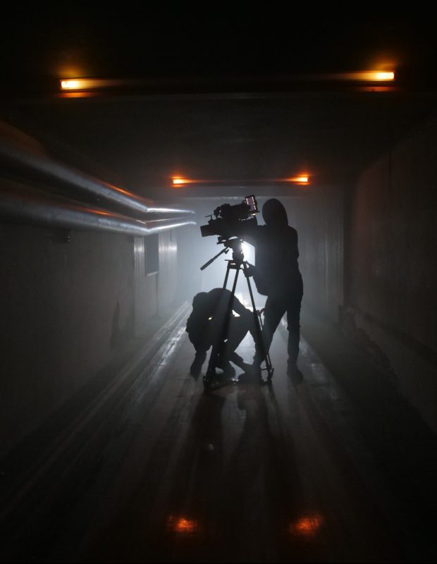 Kameramann und Assistent filmen in einem dunklen Tunnel, Silhouette gegen Licht, professionelles Filmset.