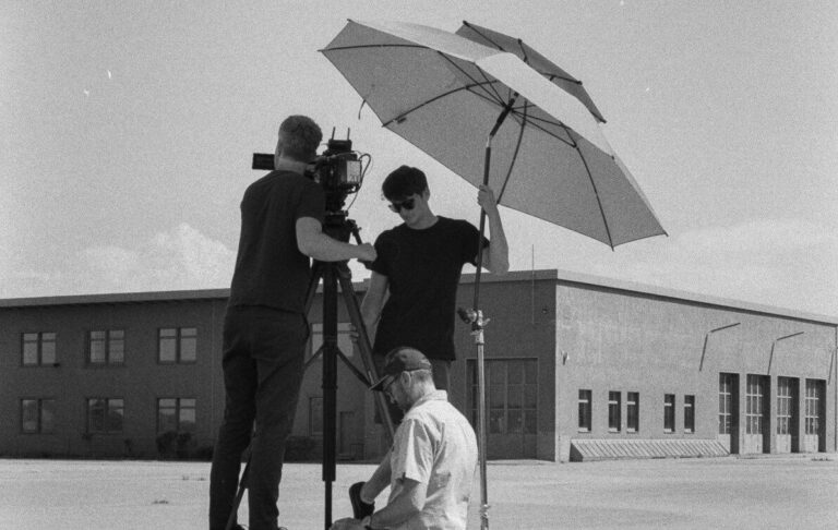 Home 40 Filmcrew bei Außenaufnahmen mit Kamera und Sonnenschirm, Schwarz-Weiß-Aufnahme, Teamarbeit am Set.