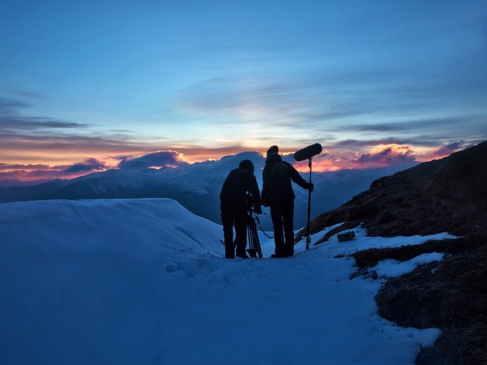 Filmcrew bei Dreharbeiten im Schnee bei Sonnenuntergang in den Bergen, symbolisiert anspruchsvolle Produktionsbedingungen.